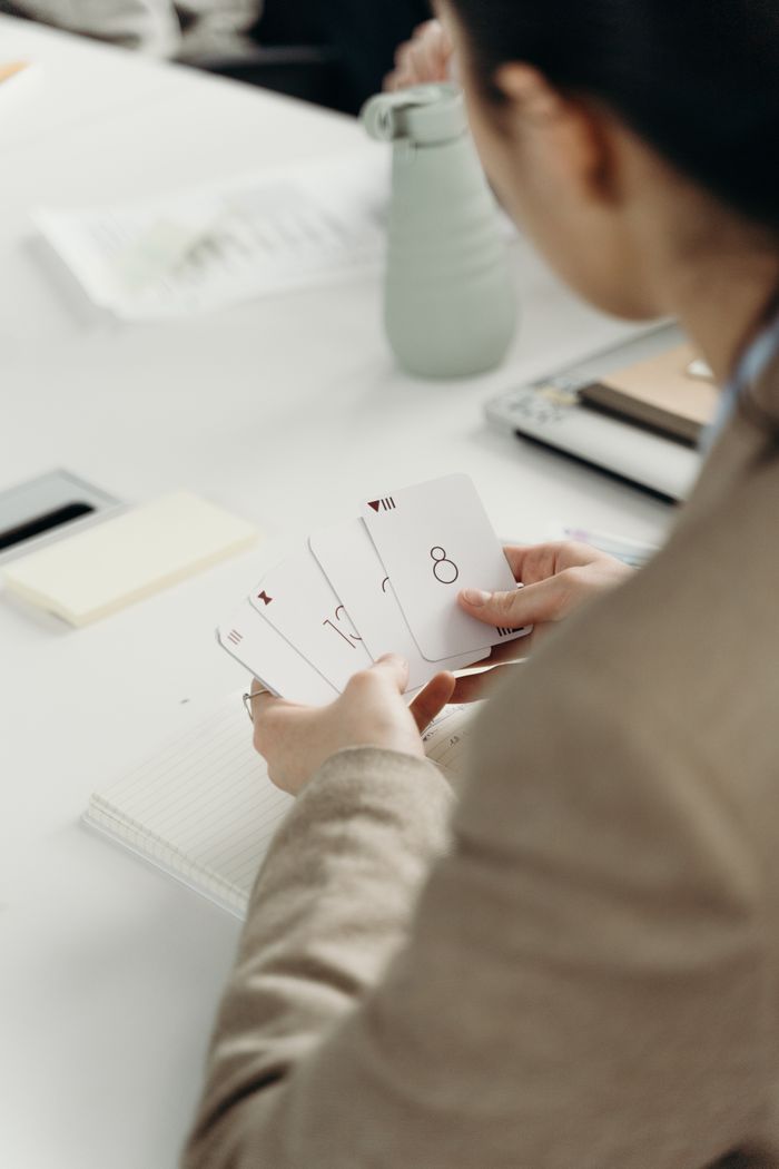 Woman holding playing cards at a white desk in an office setting, facing away from the camera.